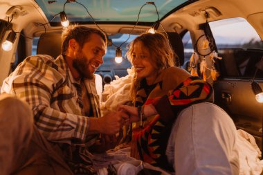 Happy young white couple smiling and lying together in car by seaside in evening