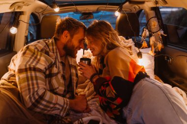 Happy young white couple smiling and lying together in car by seaside in evening