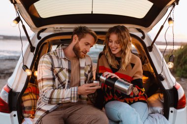 Happy young white couple smiling and drinking tea while sitting in trunk by seaside on sunset