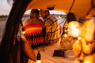 Happy young white couple smiling and playing guitar while sitting in trunk by seaside on sunset