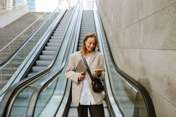 Spor Ceketli Elinde Dizüstü Bilgisayarıyla Yürüyen Merdivenin Yanında Durup Telefonunu — Stok fotoğraf