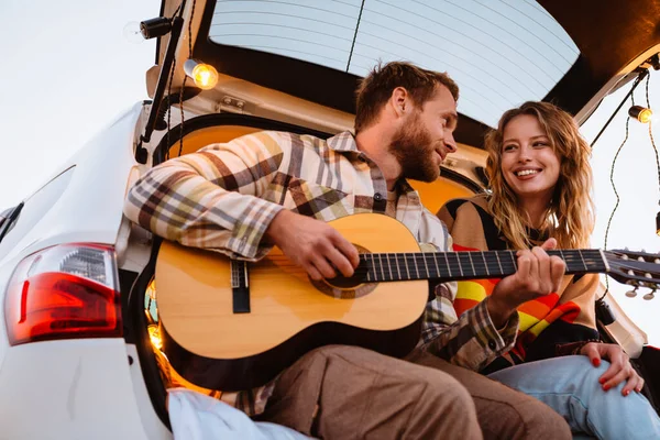 Happy young white couple smiling and playing guitar while sitting in trunk by seaside on sunset