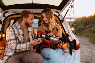 Happy young white couple smiling and drinking tea while sitting in trunk by seaside on sunset