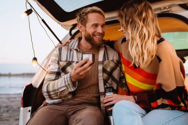 Happy young white couple smiling and drinking tea while sitting in trunk by seaside on sunset