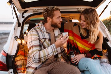 Happy young white couple smiling and drinking tea while sitting in trunk by seaside on sunset