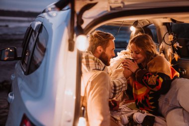 Happy young white couple smiling and lying together in car by seaside in evening
