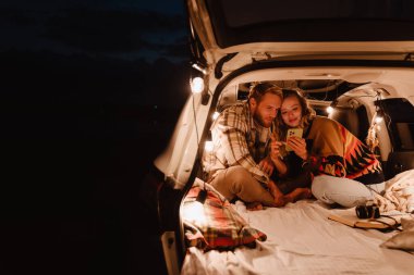 Happy young white couple using cellphone and lying together in car outdoors
