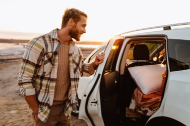 Happy young white man smiling while standing by car at seashore on sunset