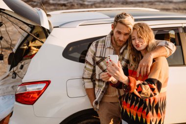 Happy young white couple taking selfie photo by car while walking at seashore on sunny day