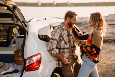 Happy young white couple standing by car while walking at seashore on sunny day