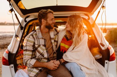 Happy young white couple smiling while sitting in trunk by seaside on sunset