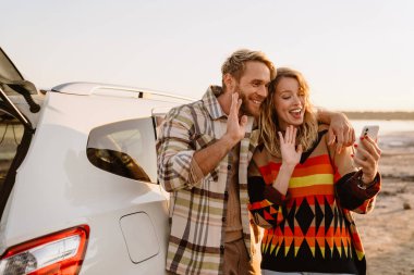 Happy young white couple taking selfie photo by car while walking at seashore on sunny day