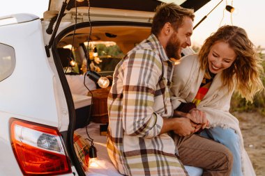Happy young white couple smiling and sitting in car trunk together outdoors