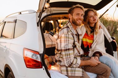 Happy young white couple smiling while sitting in trunk by seaside on sunset