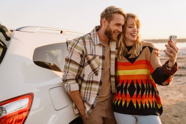 Happy young white couple taking selfie photo by car while walking at seashore on sunny day