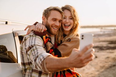 Happy young white couple taking selfie photo by car while walking at seashore on sunny day