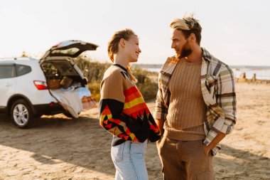 Happy young white couple smiling and looking at each other while walking by seashore