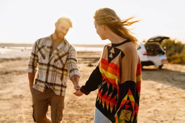 Happy young white couple smiling and holding hands while walking by seashore