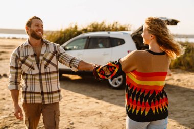 Happy young white couple smiling and holding hands while walking by seashore