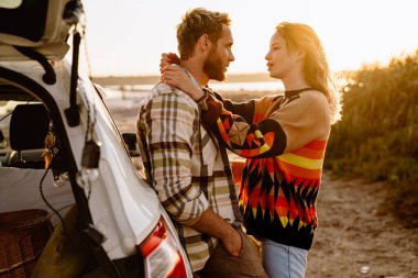 Happy young white couple standing by car while walking at seashore on sunny day