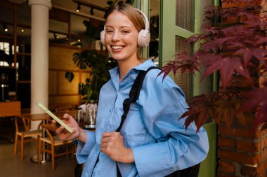 Young beautiful smiling woman in blue shirt and headphones holding her phone and looking at camera , while standing near cafe
