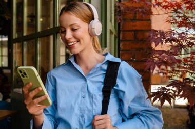 Young beautiful smiling woman in blue shirt and headphones holding and using her phone, while standing near cafe