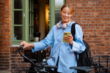 Young beautiful smiling happy stylish woman in blue shirt holding and using her phone , while standing with bicycle near brick wall outdoors