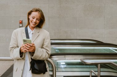 Young white woman smiling and using cellphone while standing by escalator outdoors