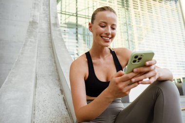 Young beautiful smiling happy sporty woman holding and using her phone , while sitting outdoors
