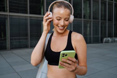 Young beautiful attractive smiling happy sporty woman in headphones with bag holding and using her phone , while standing on the street near modern building
