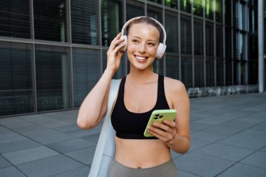 Young beautiful attractive smiling happy sporty woman in headphones with phone looking aside , while standing on the street near modern building