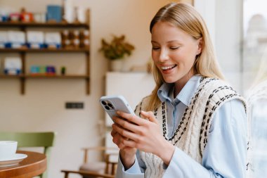 Young beautiful smiling woman in shirt and vest holding and using her phone while sitting in cozy cafe