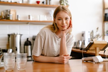 Young beautiful attractive long-haired smiling woman in a t-shirt propping her head and looking at camera , leaning on a counter, standing behind a bar in a cafe