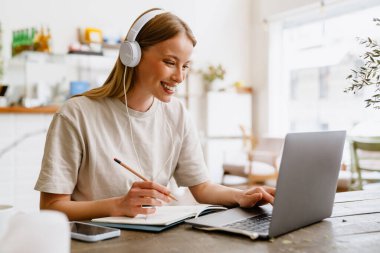Young beautiful long-haired smiling happy woman in headphones doing her homework with pencil and notebook typing on laptop while sitting in cafe