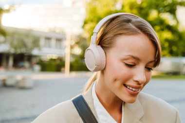 Young white woman smiling and listening to music on headphones while walking on city street