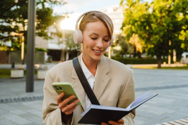 Young white woman using cellphone and note book while walking on city street