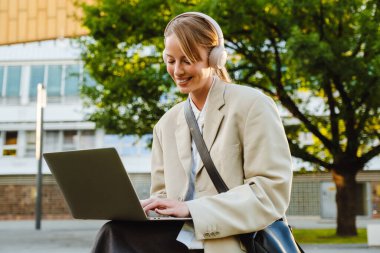 Young white woman using laptop and headphones while sitting on bench outdoors
