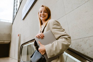 Young white woman smiling and holding laptop while standing on escalator outdoors