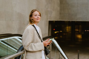 Young white woman smiling and using cellphone while standing by escalator outdoors