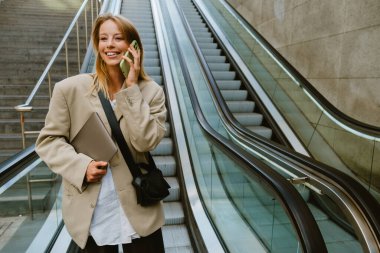 Young white woman talking on cellphone and holding laptop while going down escalator outdoors