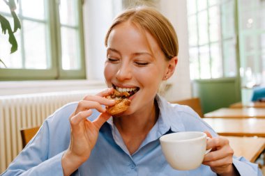 Young white businesswoman drinking coffee and eating croissant while sitting by table in cafe