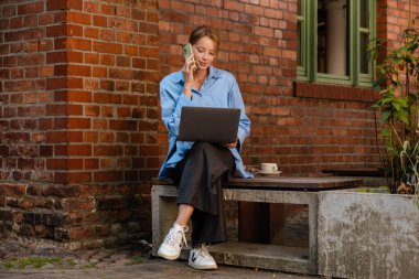Young white businesswoman talking on cellphone and using laptop while sitting in outdoor cafe