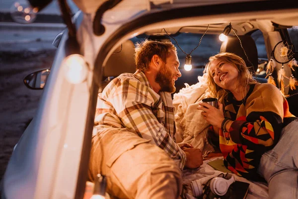 Happy young white couple smiling and lying together in car by seaside in evening