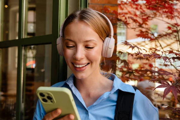 Portrait of young beautiful smiling woman in blue shirt and headphones holding and using her phone,while standing near cafe outdoors