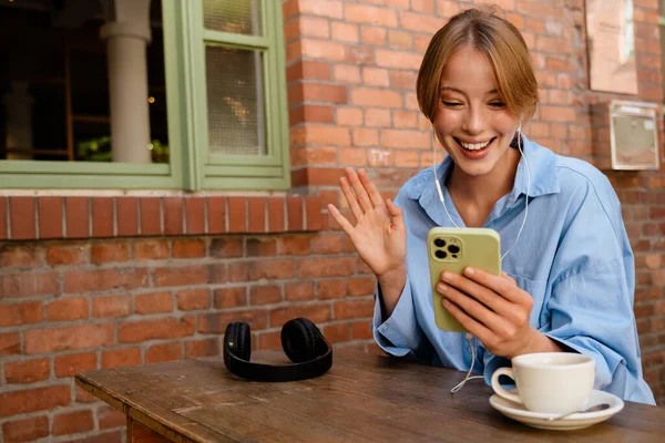Young beautiful smiling woman in headphones with phone holding video conference and waving, while sitting with cup of coffee by table near brick wall outdoors