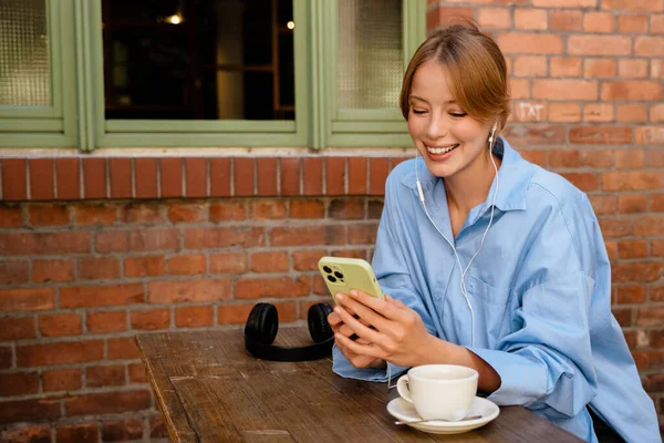 Young beautiful smiling woman in headphones holding and using her phone , while sitting with cup of coffee by table near brick wall outdoors