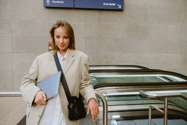 Young White Woman Smiling Holding Laptop While Standing Escalator Outdoors — Zdjęcie stockowe