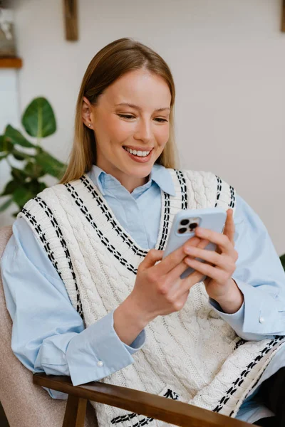 Young beautiful smiling smiling woman in shirt and vest with phone sitting in armchair