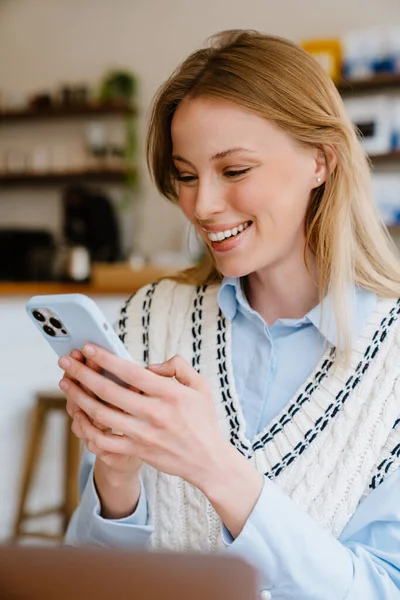 Young beautiful long-haired smiling happy woman in shirt and vest holding and using her phone, while sitting in cafe