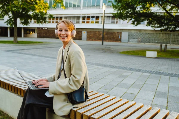 Young White Woman Using Laptop Headphones While Sitting Bench Outdoors — Stockfoto